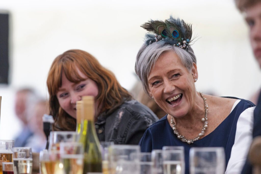 Two women at a banquet table, one with a feathered headpiece laughing heartily while the other smiles. Various drinks are placed on the table. Shot for Lydia Turner.