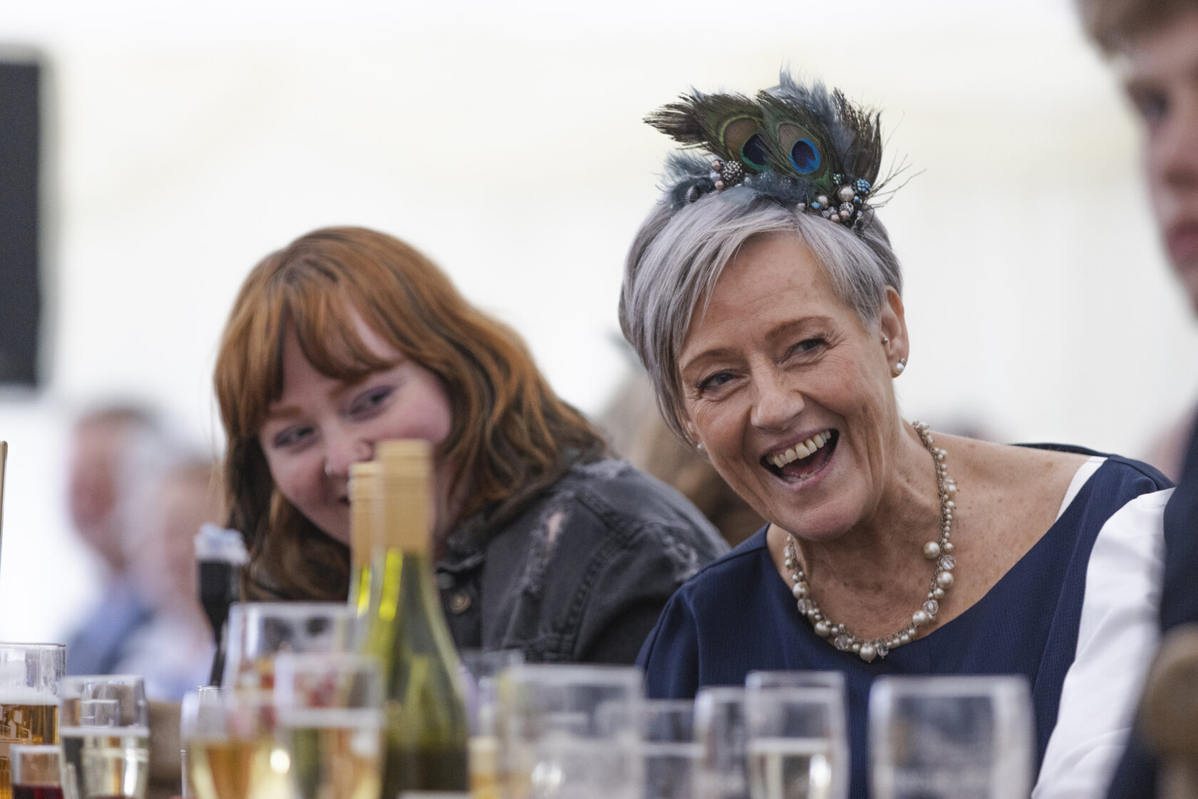 Two women at a banquet table, one with a feathered headpiece laughing heartily while the other smiles. Various drinks are placed on the table. Shot for Lydia Turner.