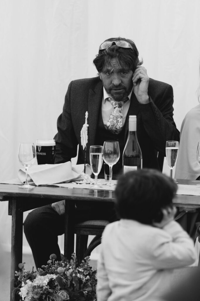 A man in a dark suit and patterned tie sits at a table with drinks, appearing deep in thought. A child is seen in the foreground, partially turned away, with colourful flowers on the table. Shot for Lydia Turner.