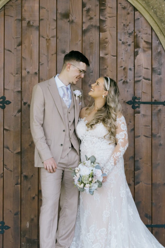 A smiling couple in wedding attire stands together in front of a wooden background. The bride wears a lace dress and holds a bouquet, while the groom is dressed in a light beige suit.