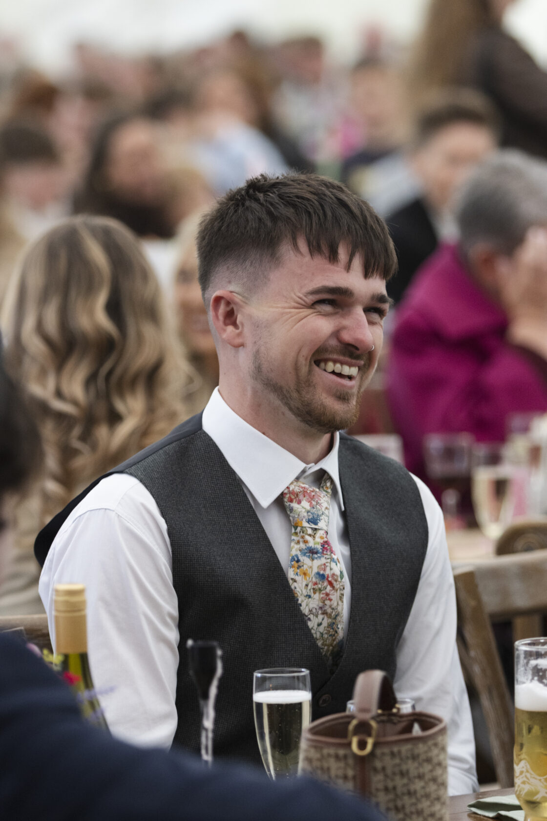 A man with short brown hair and a trimmed beard is smiling while sitting at a table during a celebration. He is dressed in a grey waistcoat and a colourful floral tie, surrounded by blurred guests in the background. Shot for Lydia Turner.