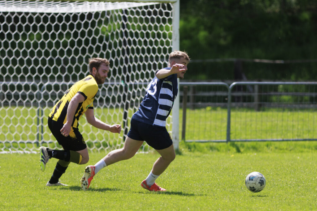 A football player in a yellow jersey is chasing after a player in a blue and white striped jersey on a grass pitch, with a goalpost in the background.