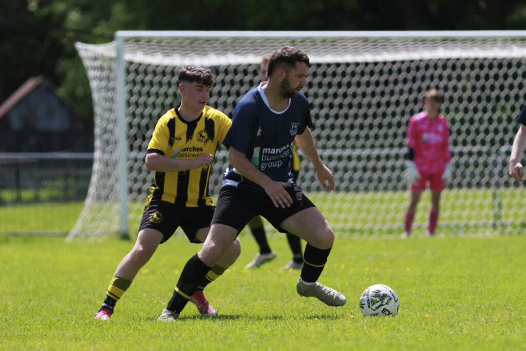 A youth football match in progress with two players competing for the ball. One player in a yellow and black striped kit is closely tracking another wearing a blue kit. A goalkeeper in a pink outfit can be seen in the background.