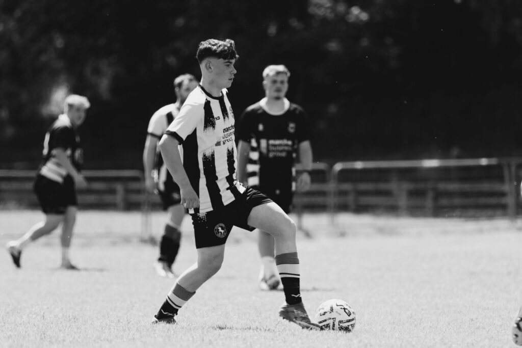A young male footballer in a black and white striped kit is poised to kick a football on a grassy pitch, with other players in the background.