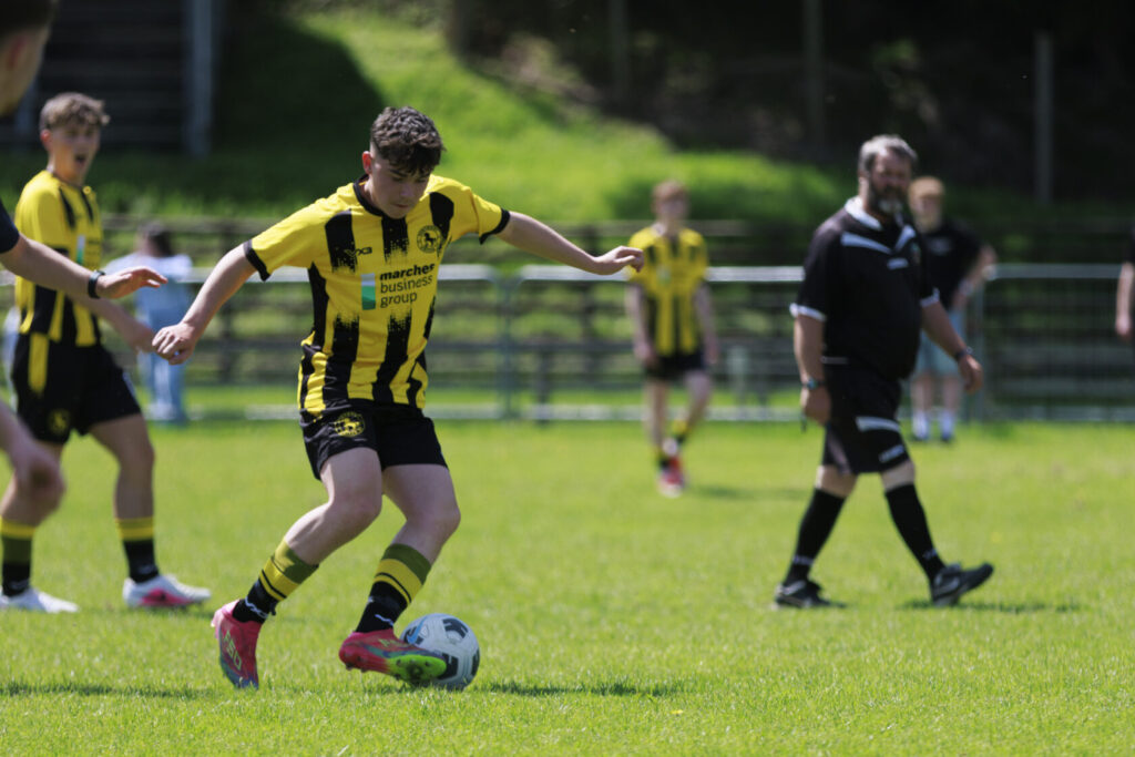 A young football player in a yellow and black striped jersey kicking a football on a bright green pitch during a youth match, with teammates and a referee in the background.