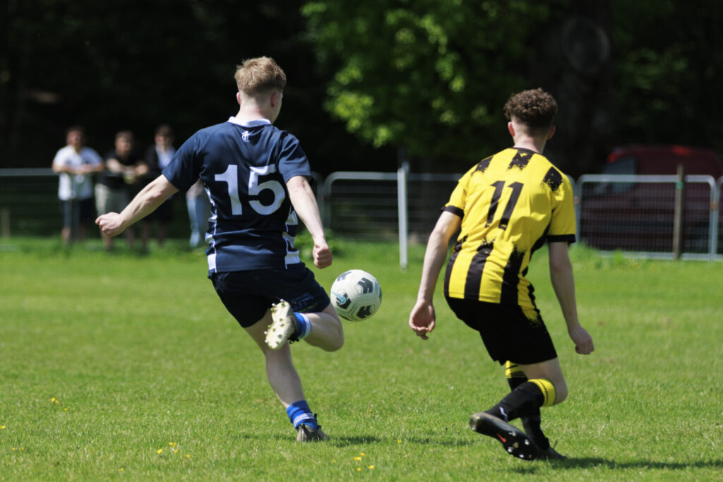 Two young football players competing on the pitch. One player in a dark blue kit, number 15, is striking a ball, while the other in a yellow and black striped kit, number 11, is closely following behind.