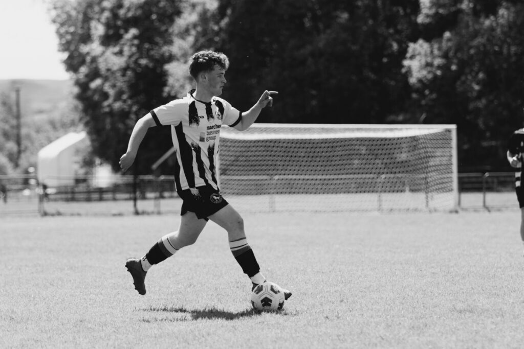 A young male football player wearing a black and white striped jersey and shorts, running on a grassy field with a football at his feet. A goal post can be seen in the background.
