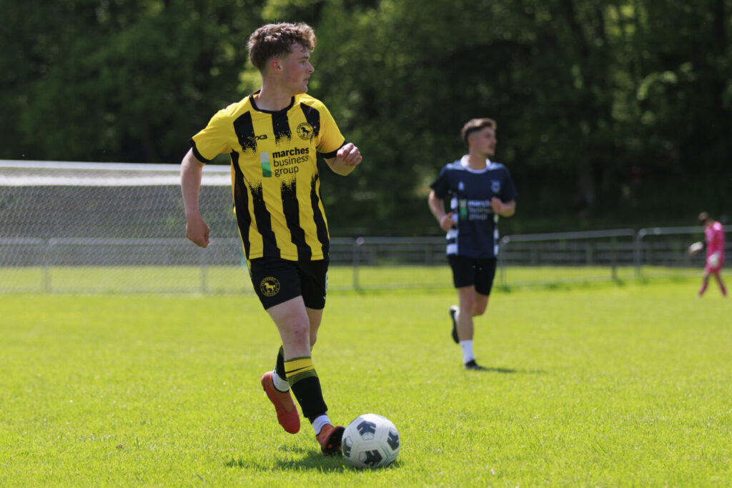 A young football player wearing a yellow and black striped jersey dribbles a football on a lush green pitch during a match, with teammates and opponents in the background.