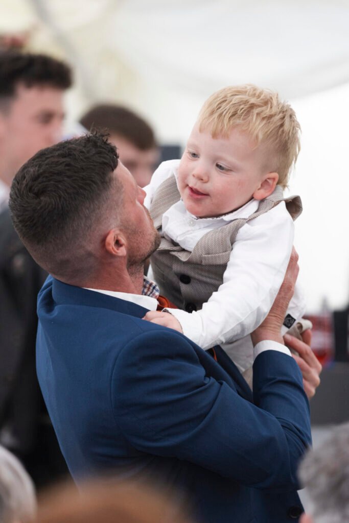 A man in a blue suit lifts a small boy, who is smiling and wearing a grey vest and white shirt. The background shows people mingling at a festive event. Shot for Lydia Turner.
