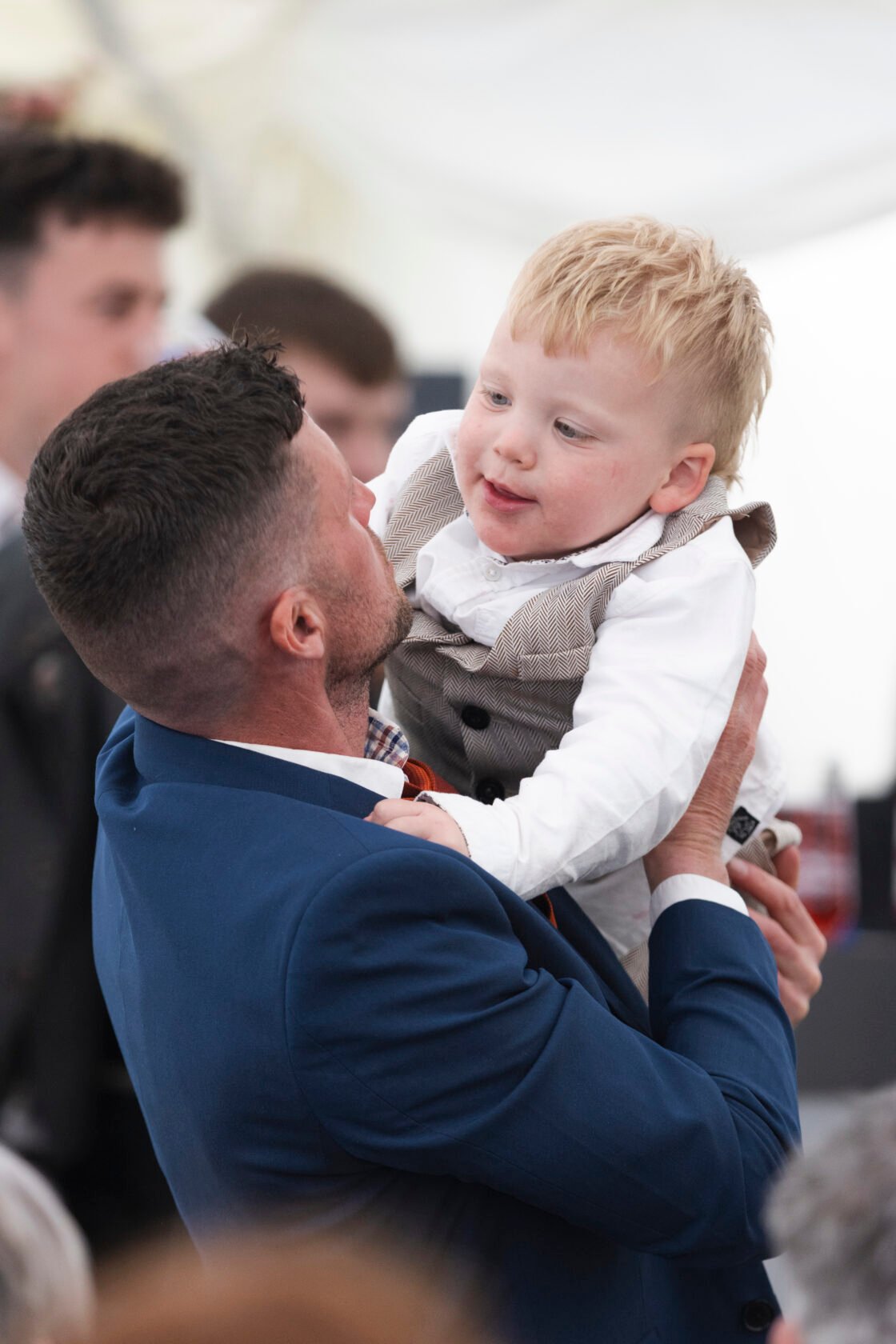 Father and Child Bonding at Event A man in a blue suit lifts a small boy, who is smiling and wearing a grey vest and white shirt. The background shows people mingling at a festive event. Shot for Lydia Turner.