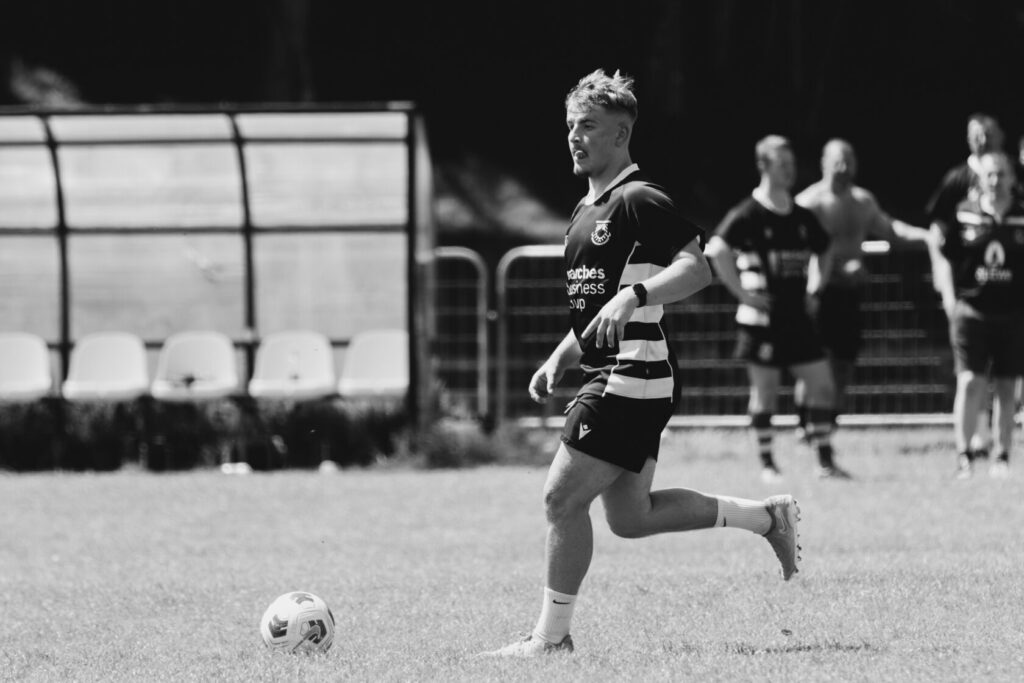 A rugby player in a black and white kit runs across a grassy field during a training session, with a ball nearby and teammates visible in the background.