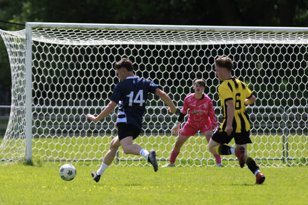 A player in a blue jersey with the number 14 attempts to score while a goalkeeper in pink and another player in yellow with the number 6 react during a youth football match.