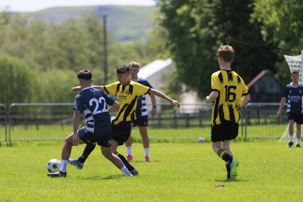 A youth football match featuring players in blue and yellow kits on a grassy pitch under sunny skies. One player in a blue kit attempts to evade an opponent in yellow while a third player observes.