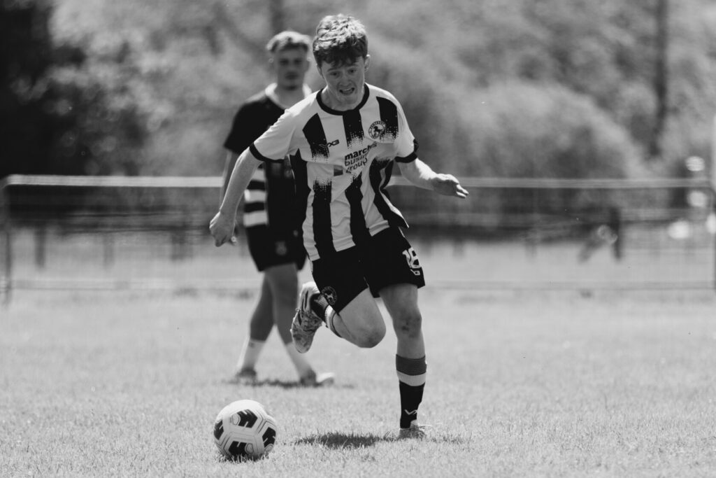 A young football player in a black and white striped jersey kicking a football on a grassy pitch, with another player in the background.