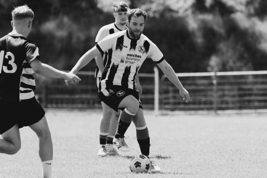 A black and white photograph of a football player in a striped jersey kicking a ball during a match. Another player in the background is reaching out to assist.