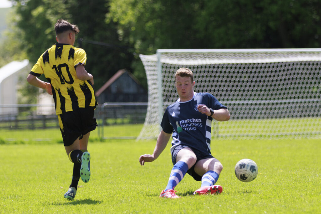 A youth football player in a yellow and black striped kit dribbles past a sliding opponent in a blue kit during a sunny day match on a grassy pitch.