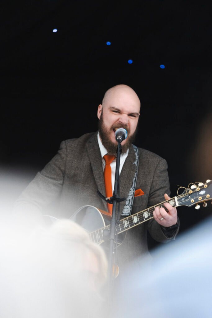 A bearded musician on stage passionately singing into a microphone while playing an acoustic guitar, dressed in a brown suit and orange tie. Shot for Lydia Turner.