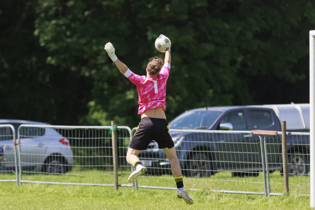 A goalkeeper in a pink jersey jumps to catch a football during a match, with a fence and parked cars in the background.