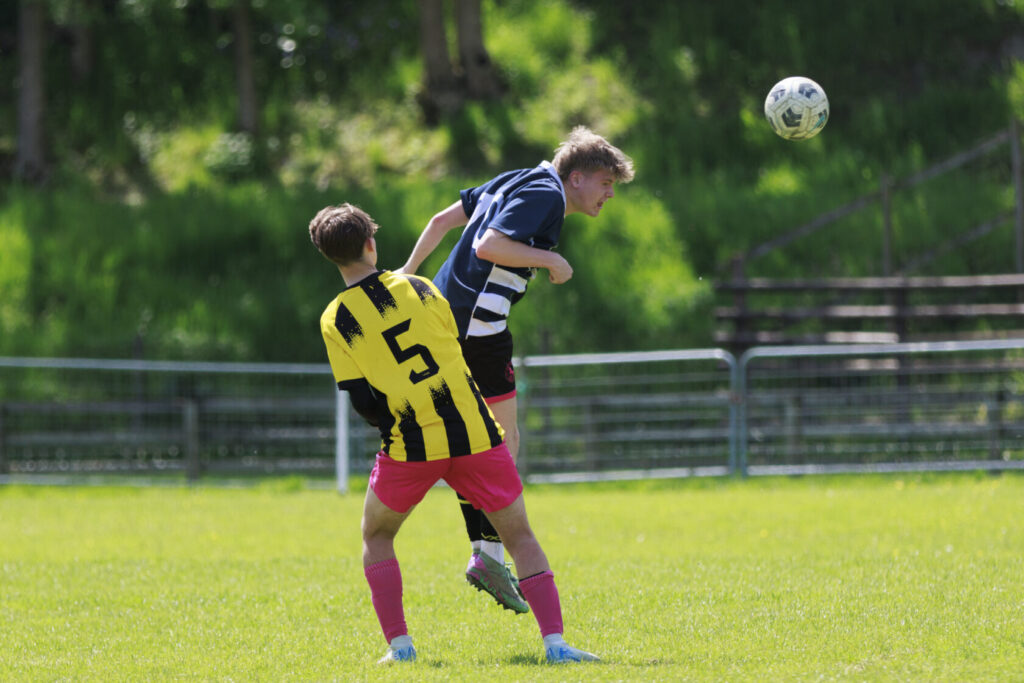 Two young male football players competing for the ball during a match. One player in a black and white striped shirt jumps to head the ball, while the other player in a yellow and black striped shirt stands behind, wearing pink shorts.