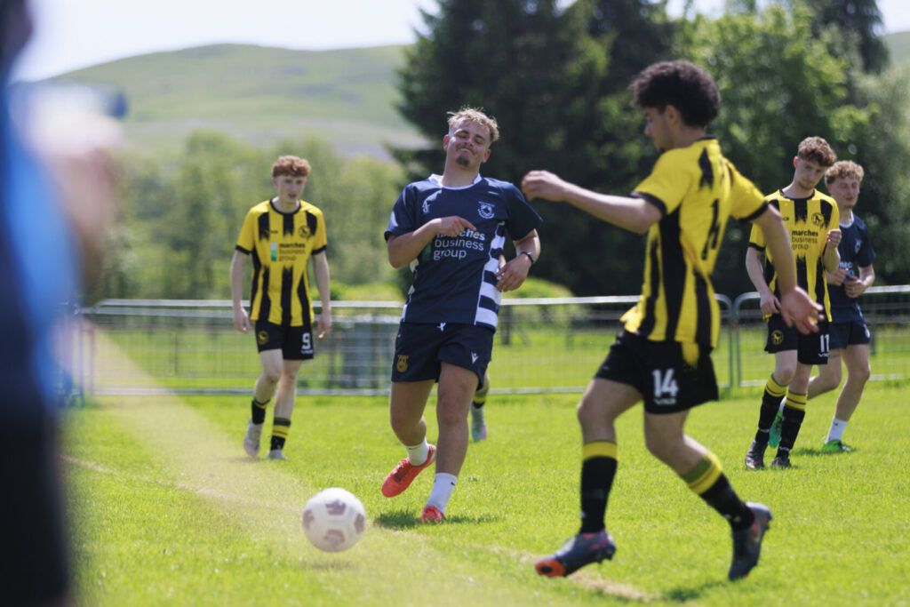 A youth football match in progress with players in blue and yellow kits on a grassy field. One player is about to kick the ball, while others are running nearby.