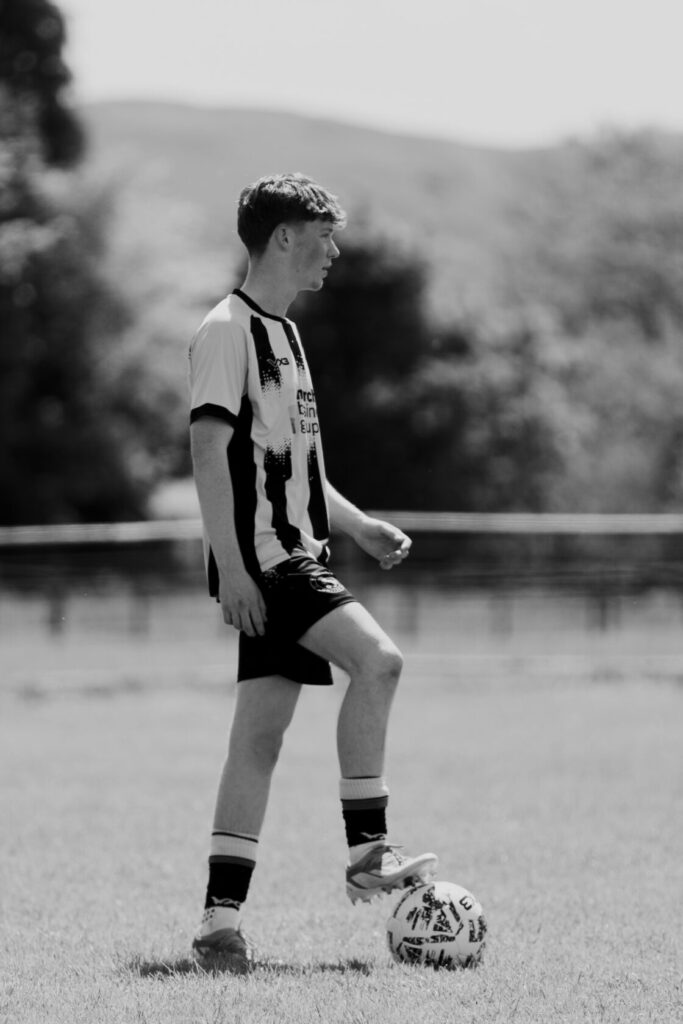 A young male footballer with short hair stands on a grassy pitch, wearing a black and white striped jersey, black shorts, and sports shoes, poised to kick a football on the ground.
