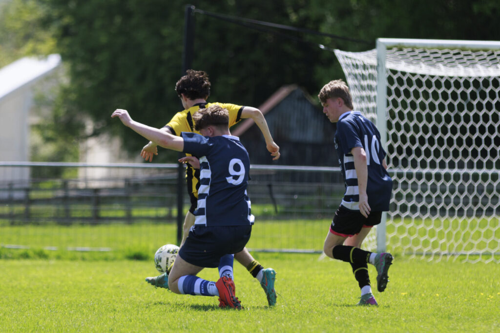 A youth football match where players in navy and yellow kits compete for the ball. One player is on the ground reaching for the ball as another player runs past.