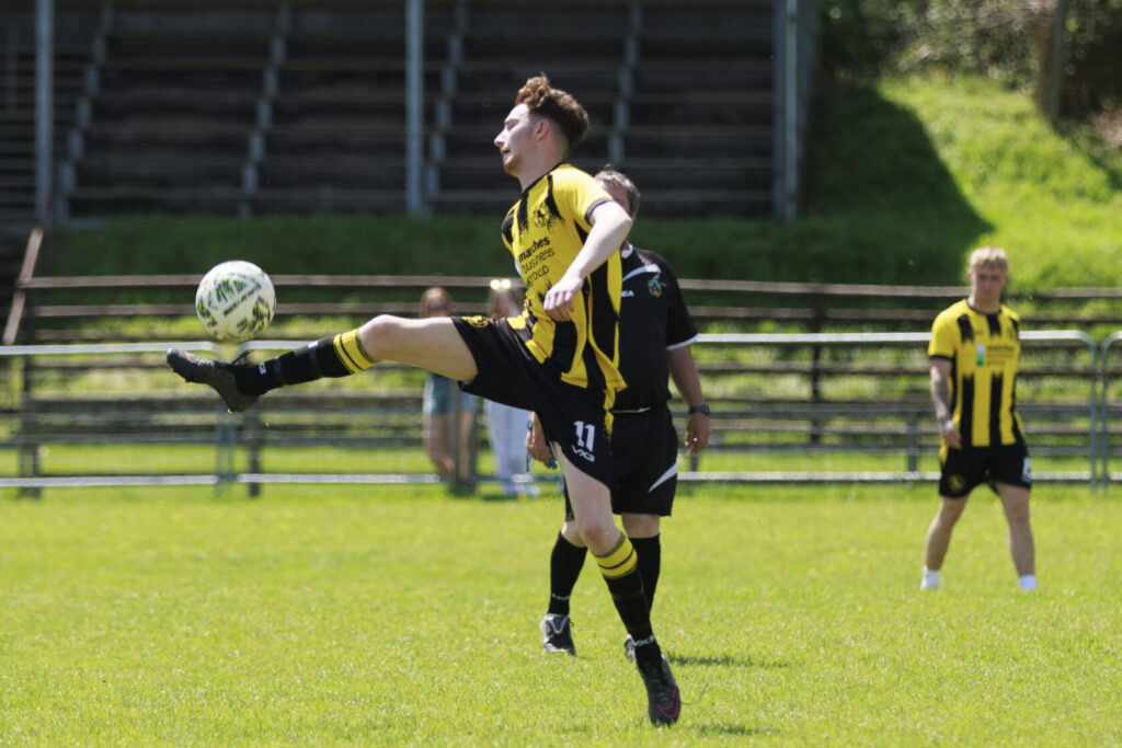 A football player in a yellow and black jersey performs a high kick to strike the ball while two other players look on in the background.
