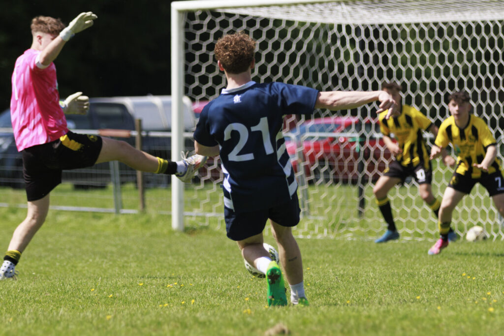 A football player in a blue jersey with the number 21 attempts to score against a goalkeeper in a pink jersey while two players in yellow jerseys watch from the background.