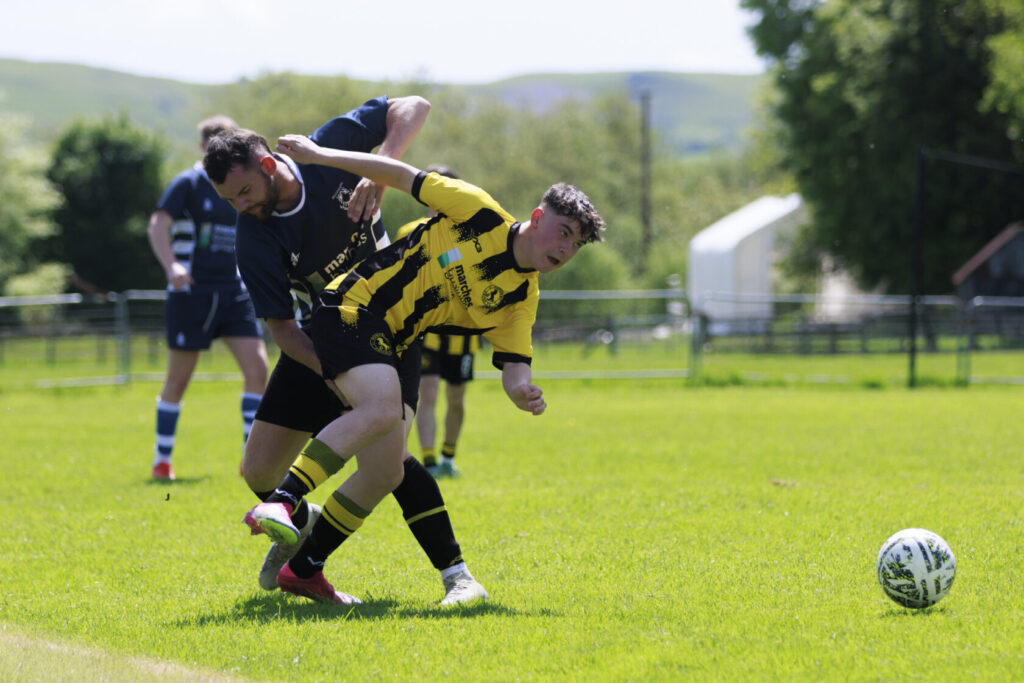 Two football players engaged in a competitive play, one in a yellow jersey and the other in a dark blue jersey, vying for the ball on a sunny day.