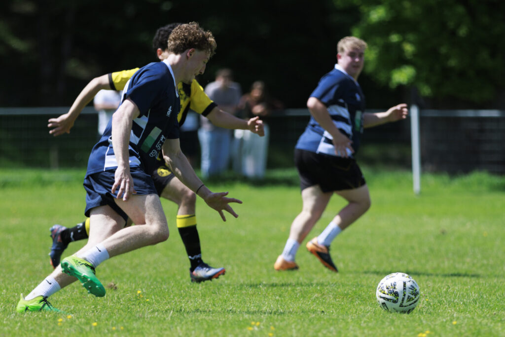 Two young footballers competing on a green pitch, one player dribbling the ball while another runs in the background, wearing contrasting team kits.