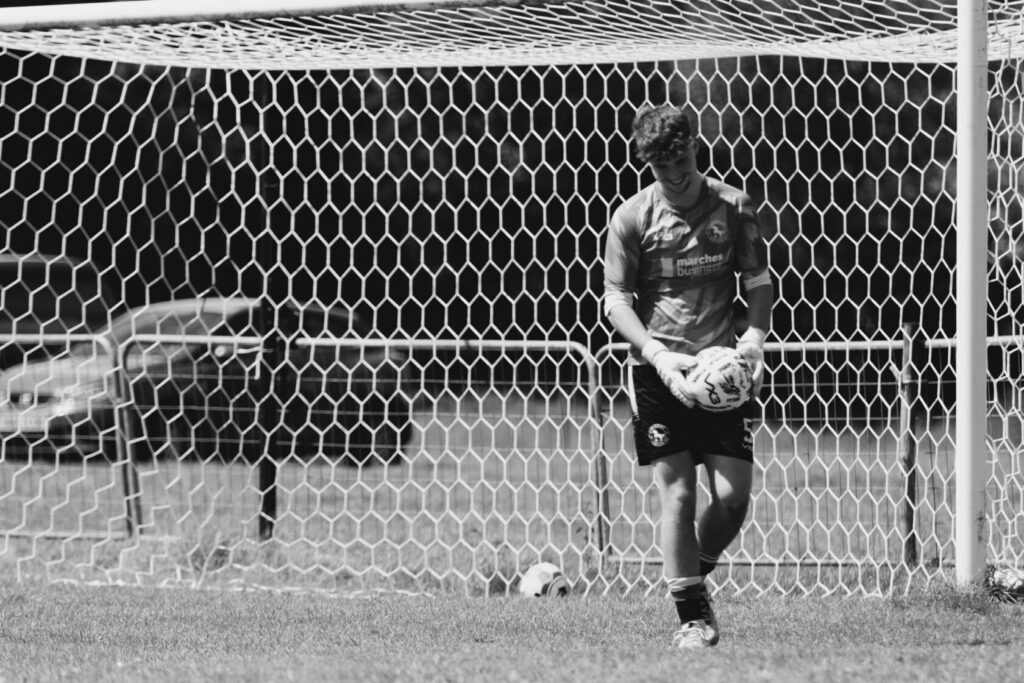 A young male goalkeeper wearing a grey jersey and black shorts stands in front of a football goal, holding a football and smiling, with a blurred background of cars and grass.