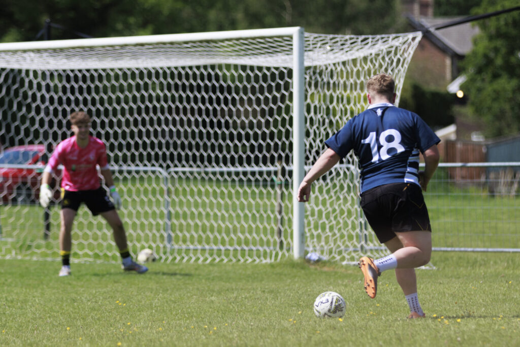 A young football player in a blue jersey, number 18, steps up to take a shot at goal while a goalkeeper in a pink jersey prepares to defend, with a goalpost and grassy field in the background.