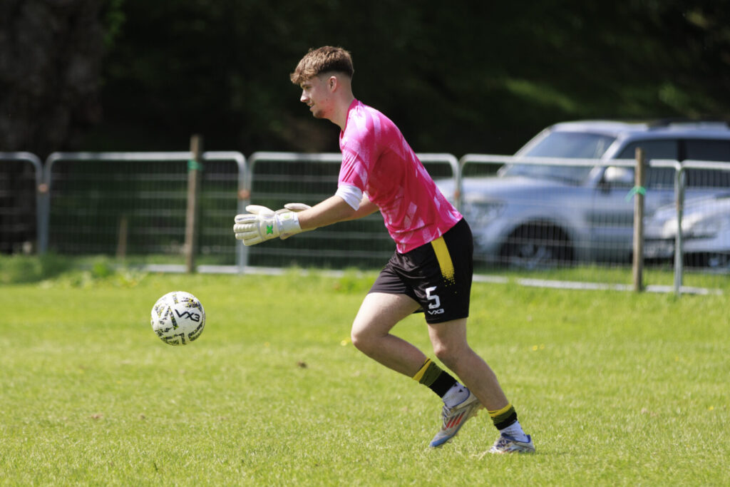 A young male goalkeeper in a bright pink jersey and gloves prepares to catch a football during a training session on a grassy field.