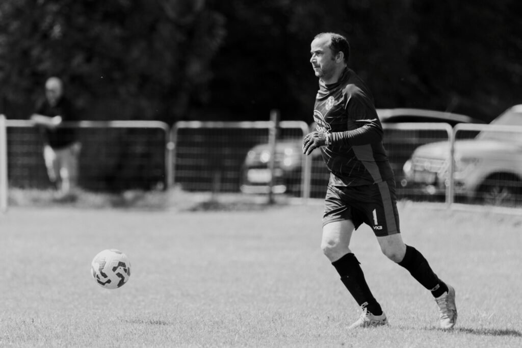 A male goalkeeper in a black and grey football kit prepares to play on a grassy pitch, with a football hovering nearby and spectators in the background.