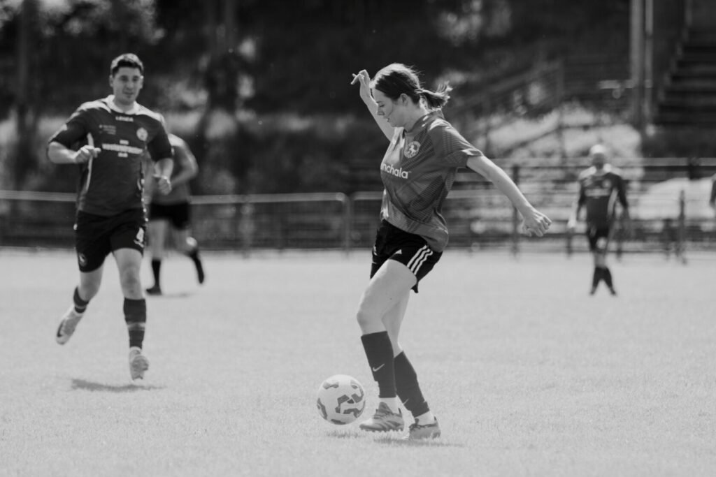A female football player in a striped jersey skillfully dribbles a ball on a lush green pitch, with another player in the background.