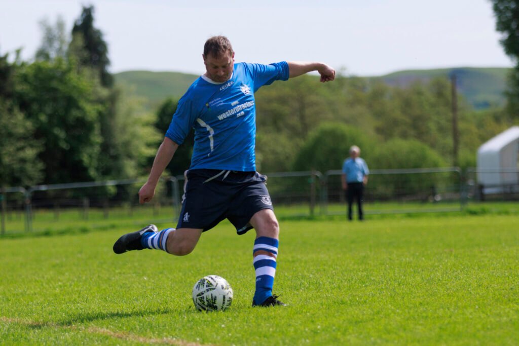 A player from Pen-y-Bont in a blue jersey and black shorts takes a shot at a football during a fundraising match for Macmillan Cancer Support and The British Heart Foundation, with a green field and trees in the background.