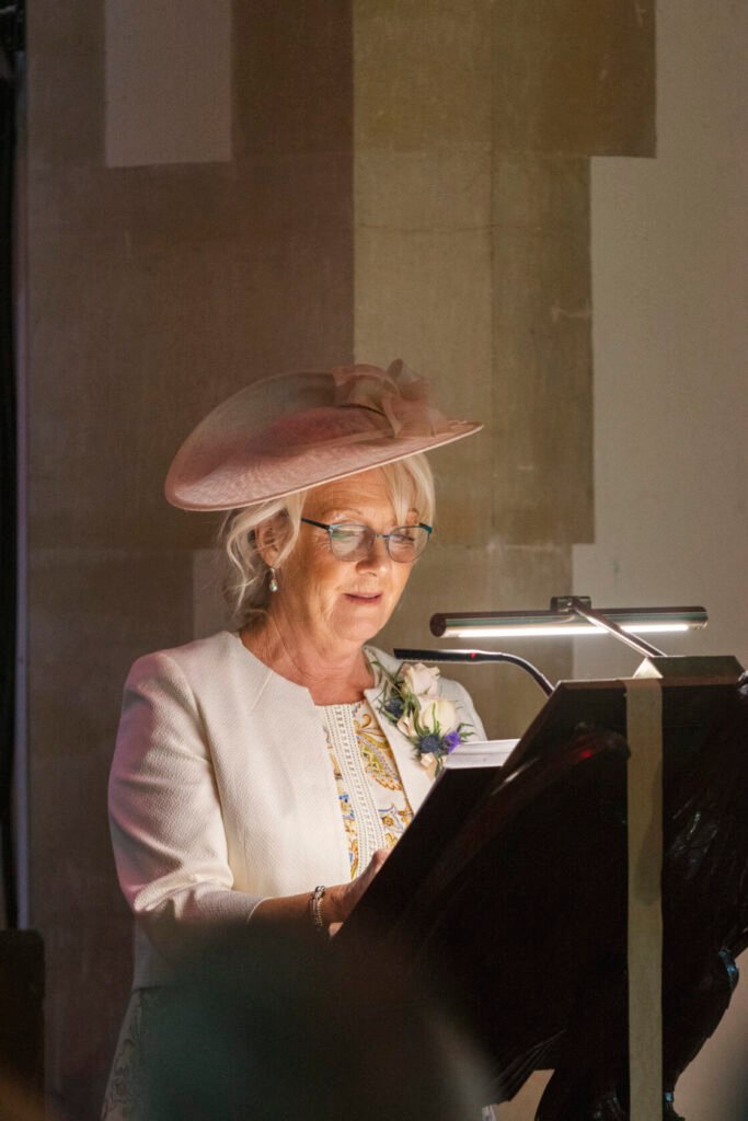 A woman with grey hair and glasses, wearing a stylish hat and a white jacket, reading from a lectern at an event. Shot for Lydia Turner.