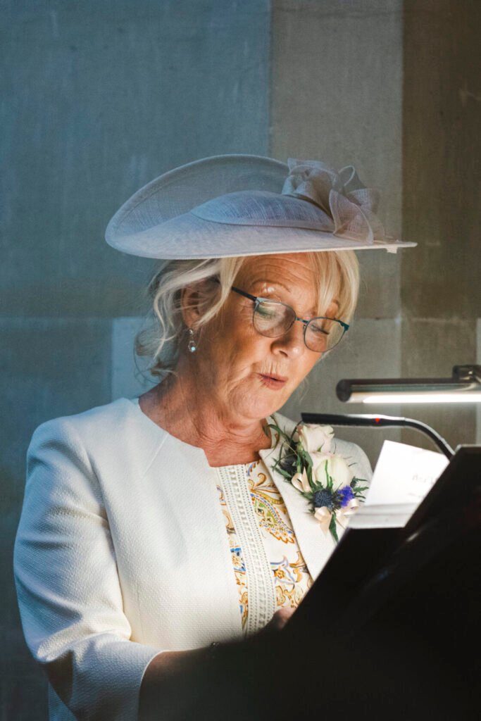 A woman with blonde hair in glasses, wearing a white jacket and a large stylish hat, reading from a music stand during a ceremony. She has a floral decoration on her lapel and appears to be deeply engaged in her reading. Shot for Lydia Turner.