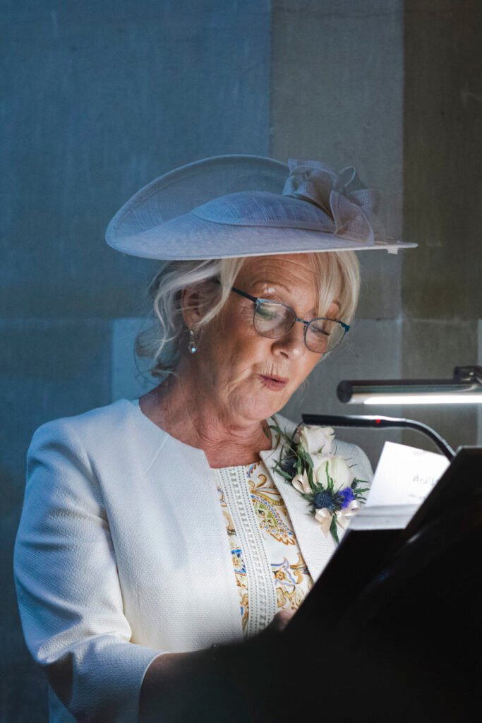 A woman with blonde hair and glasses, wearing a stylish white hat and jacket, reading from a lectern. She has a flower corsage and appears to be giving a speech at a formal event. Shot for Lydia Turner.