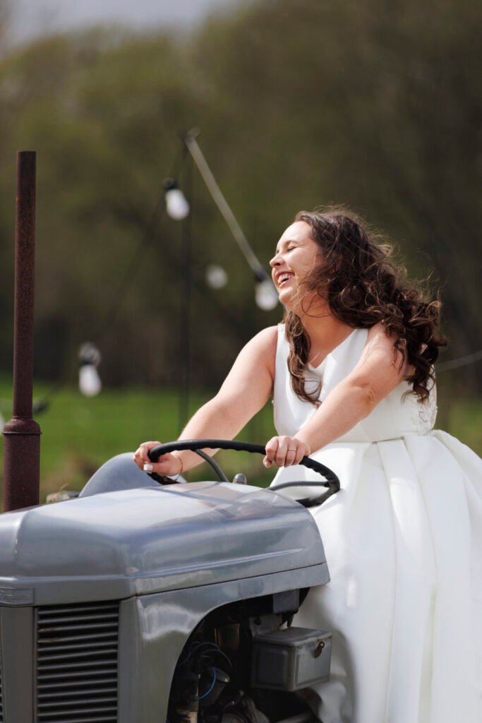 A joyful bride in a white wedding dress, laughing while driving a grey tractor, surrounded by lush green fields and decorative lights in the background. Shot for Lydia Turner.