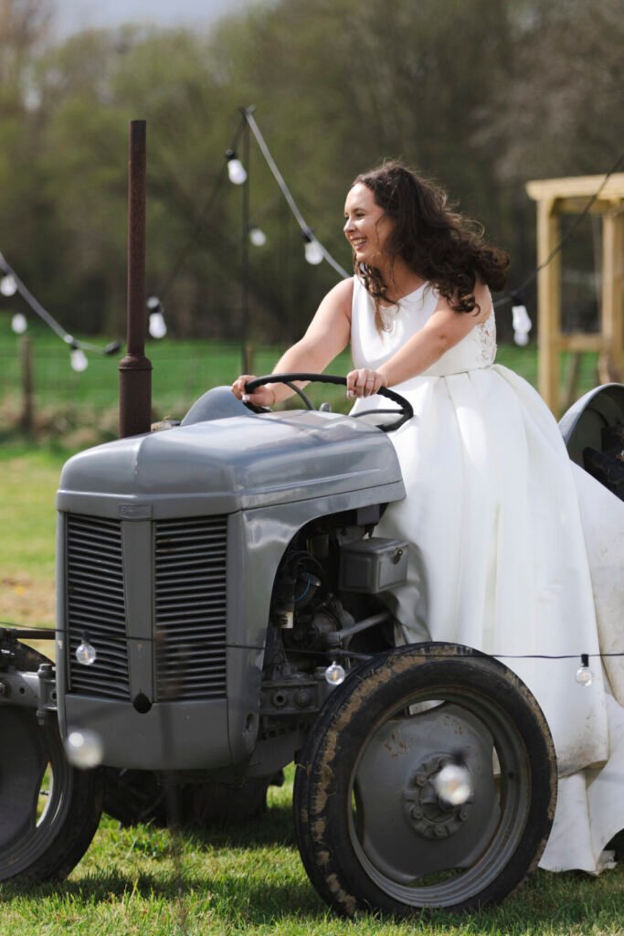 A bride in a white wedding dress laughing while sitting on a vintage grey tractor in a field, with decorative lights in the background. Shot for Lydia Turner.