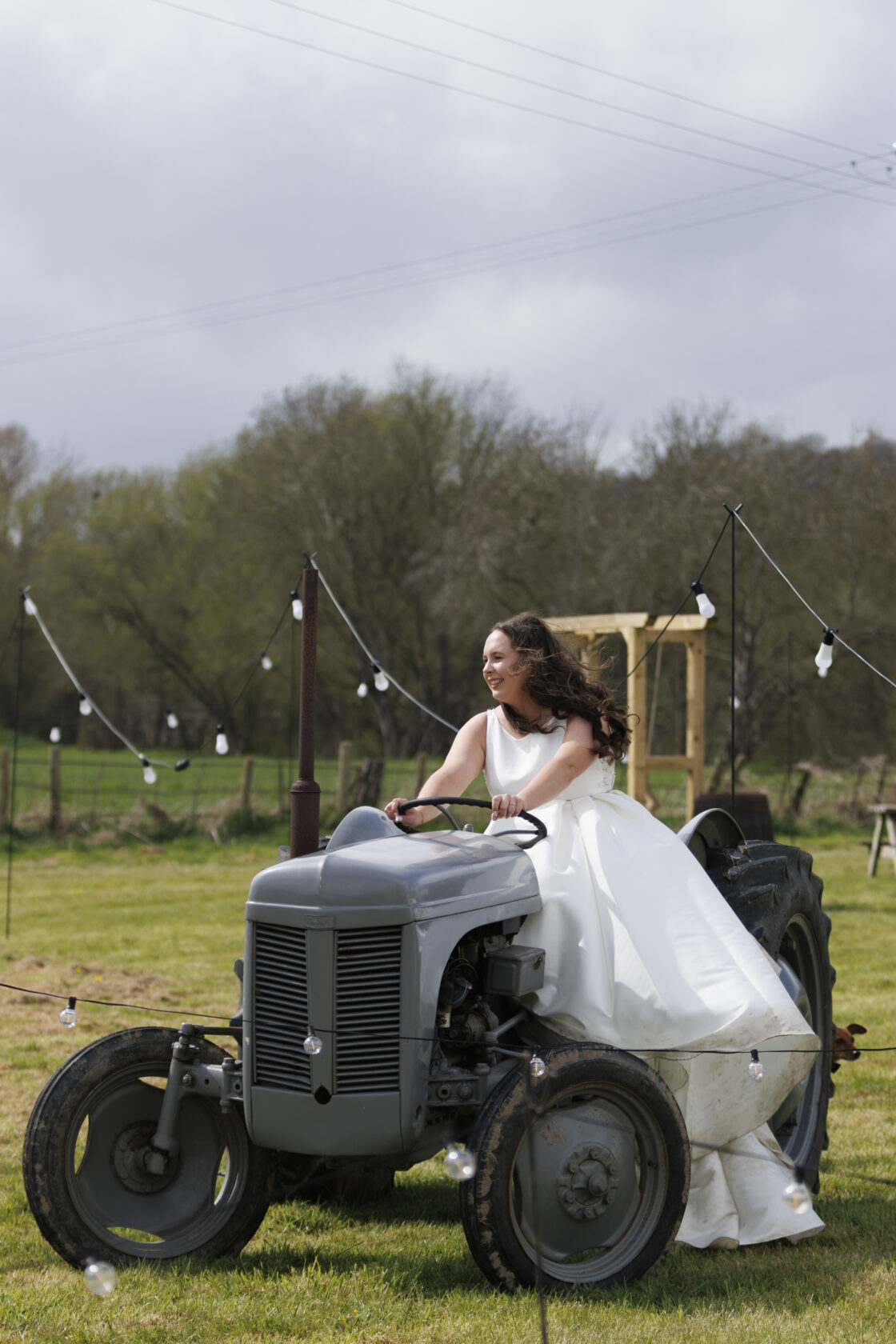 A bride in a white gown joyfully driving a vintage grey tractor on a grassy field, surrounded by decorative string lights and trees in the background. Shot for Lydia Turner.