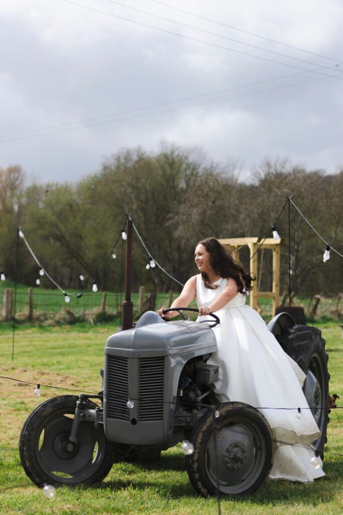 A smiling bride with long curly hair in a white wedding gown joyfully riding a vintage grey tractor. The scene is set outdoors with green fields and decorative light bulbs in the background. Shot for Lydia Turner.
