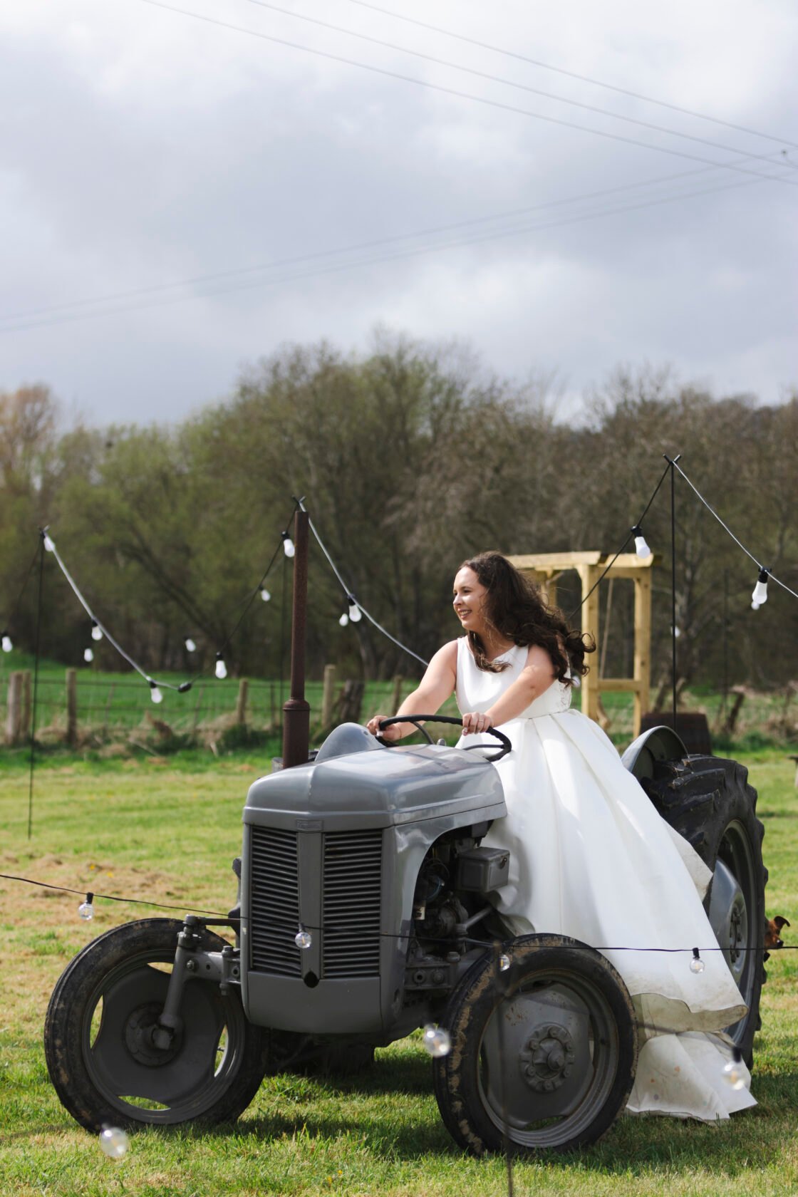 Bride Riding a Vintage Tractor at Outdoor Wedding A smiling bride with long curly hair in a white wedding gown joyfully riding a vintage grey tractor. The scene is set outdoors with green fields and decorative light bulbs in the background. Shot for Lydia Turner.
