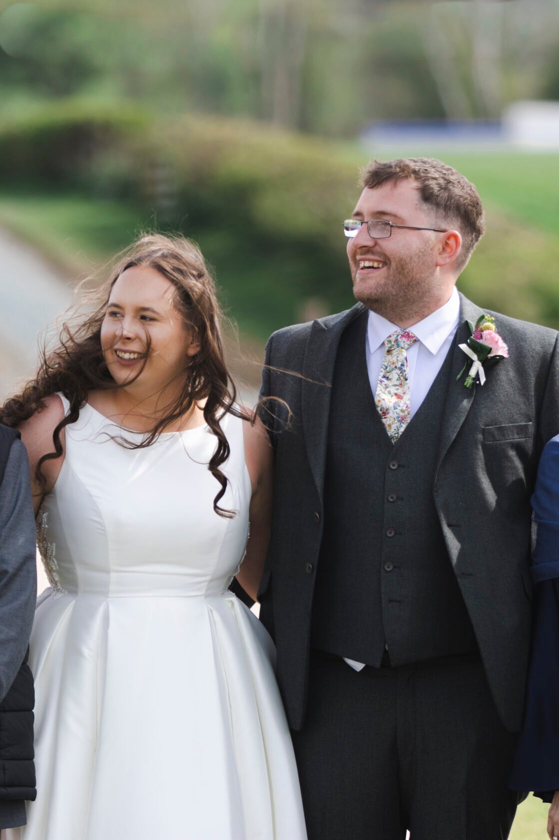 Joyful Wedding Moment A bride in a white dress smiles warmly while standing next to a man in a grey suit, both enjoying a sunny day. Shot for Lydia Turner.