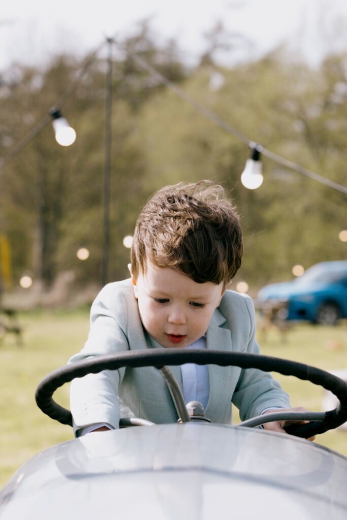 A young boy in a light blue suit leaning over the steering wheel of a classic toy car, focused and engaged. Soft background with greenery and hanging lights. Shot for Lydia Turner.