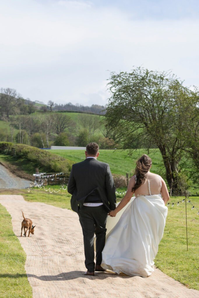 A bride in a flowing white gown and a groom in a grey suit walk hand-in-hand along a rustic path, with a dog wandering nearby and lush green hills in the background. Shot for Lydia Turner.