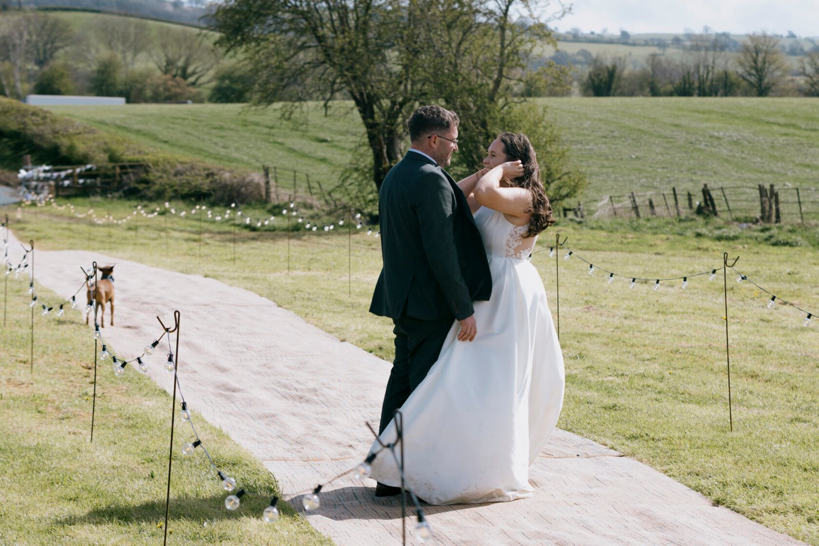 Romantic Outdoor Wedding Moment A couple sharing a tender moment during their outdoor wedding ceremony on a grassy pathway, adorned with string lights and a dog in the background. Shot for Lydia Turner