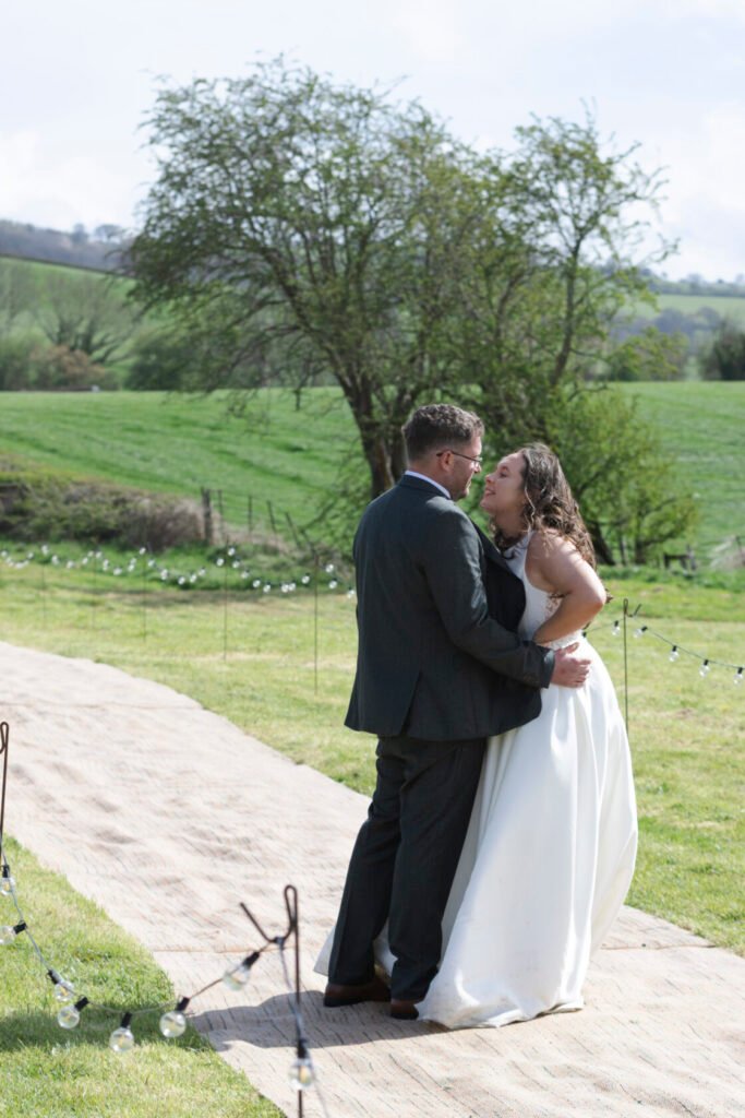 A joyful bride and groom share an intimate moment on their wedding day, surrounded by lush green fields and string lights. Shot for Lydia Turner.