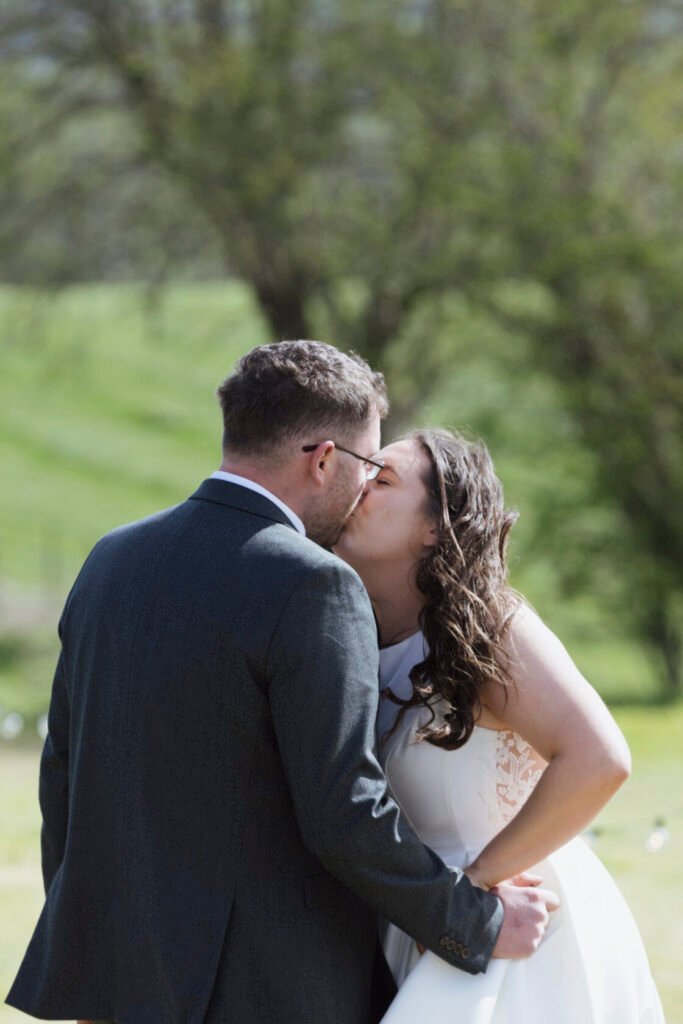 A bride and groom share a loving kiss outdoors, surrounded by lush greenery. The bride is wearing a white dress with delicate lace detailing, and the groom is in a grey suit. Shot for Lydia Turner.